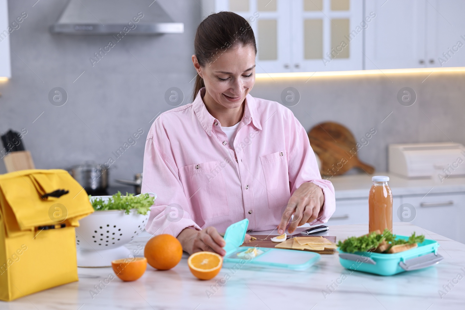 Photo of Smiling woman making snacks for school lunch box at white marble table in kitchen