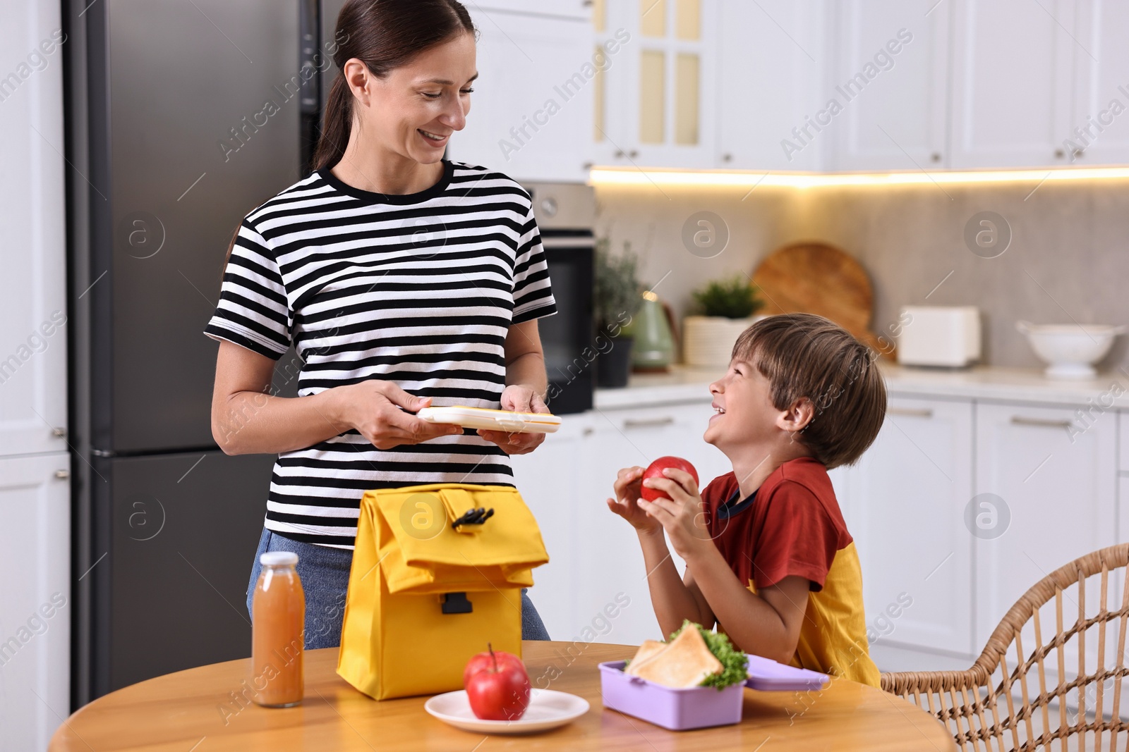 Photo of Smiling mother and her cute son preparing school lunch box with healthy food at wooden table in kitchen