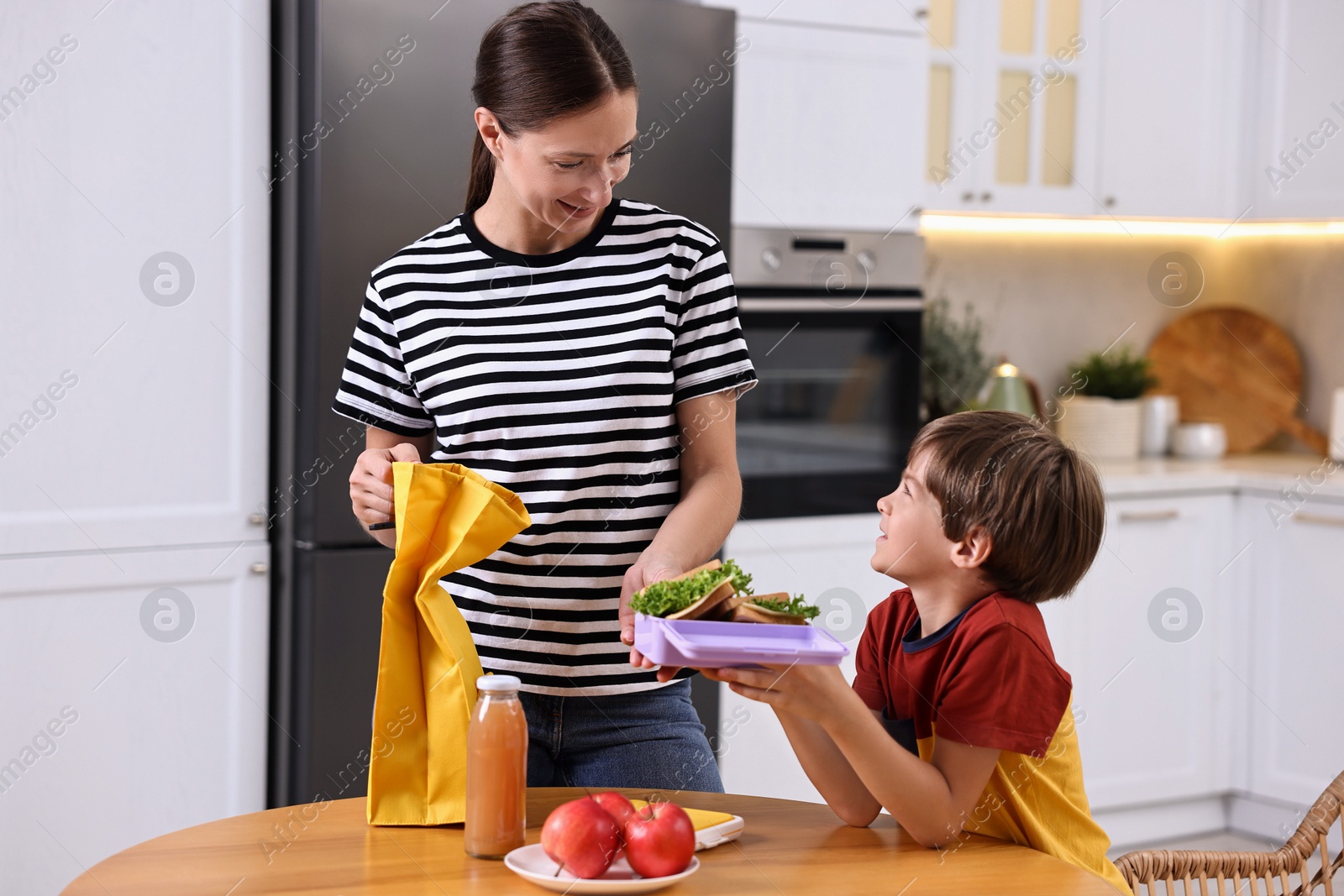 Photo of Smiling mother and her cute son preparing school lunch box with healthy food at wooden table in kitchen