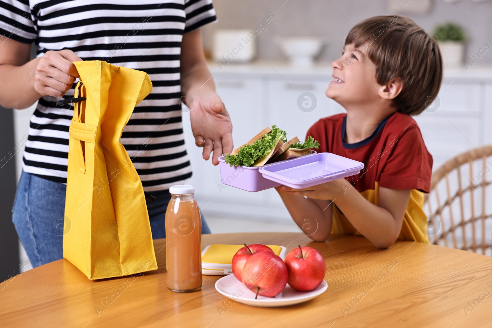 Photo of Mother and her cute son preparing school lunch box with healthy food at wooden table in kitchen, closeup