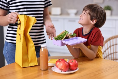 Photo of Mother and her cute son preparing school lunch box with healthy food at wooden table in kitchen, closeup