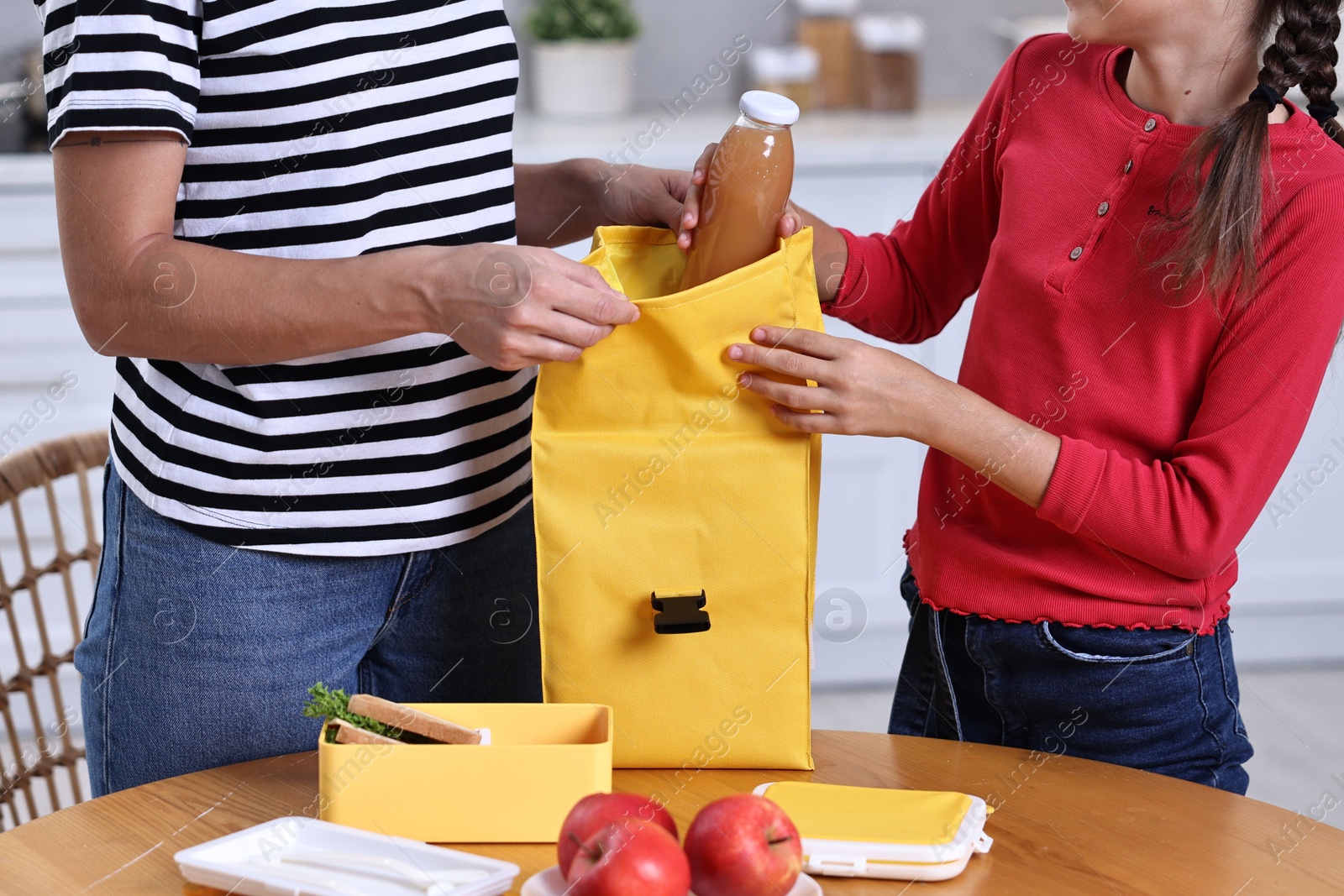 Photo of Mother putting bottle of drink into daughter`s lunch bag at wooden table in kitchen, closeup. Preparing for school