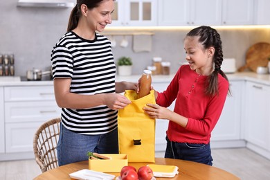 Photo of Mother with her daughter putting bottle of drink into lunch bag at wooden table in kitchen. Preparing for school