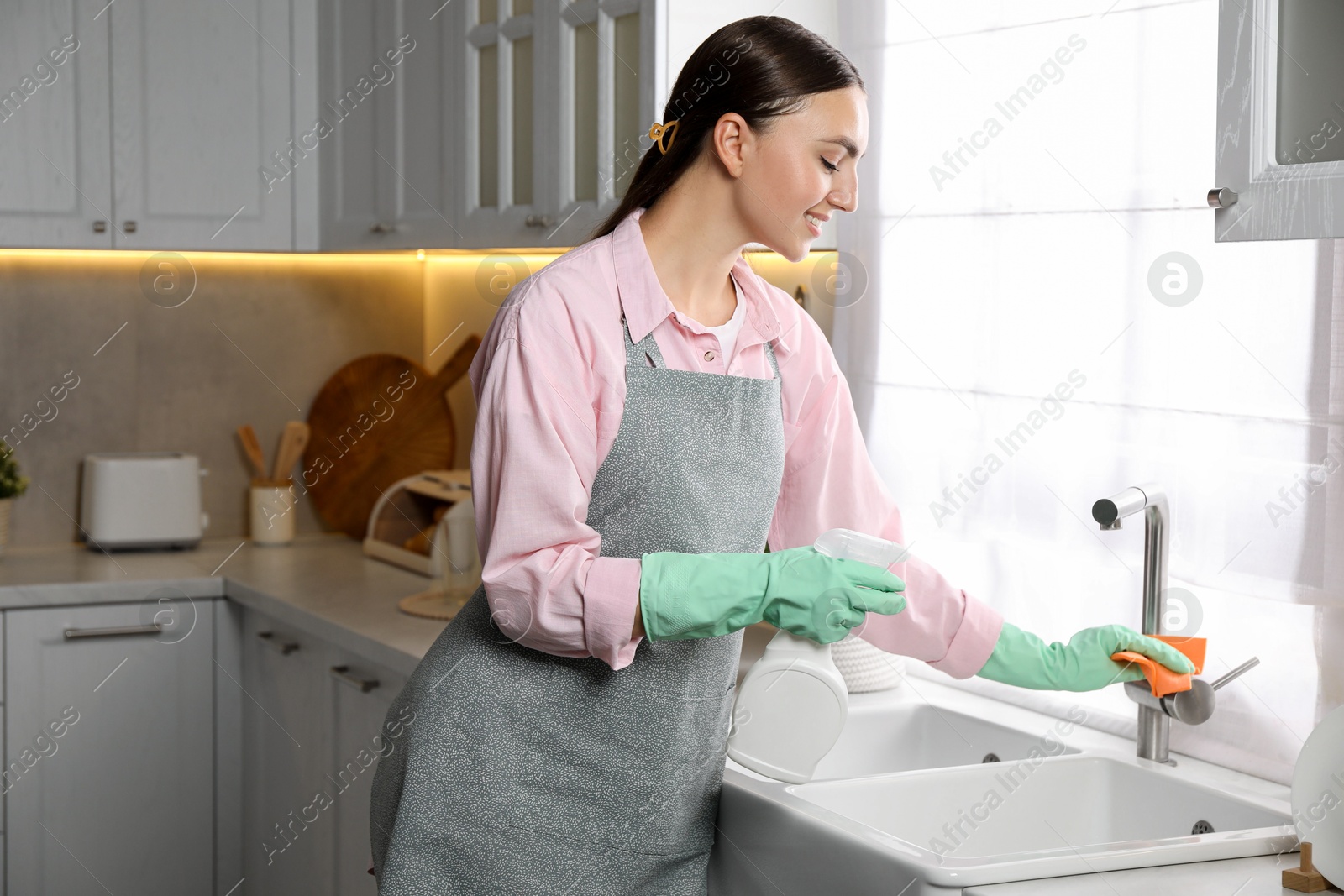 Photo of Beautiful young woman cleaning tap with spray bottle of detergent and napkin in kitchen