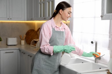 Beautiful young woman cleaning tap with spray bottle of detergent and napkin in kitchen