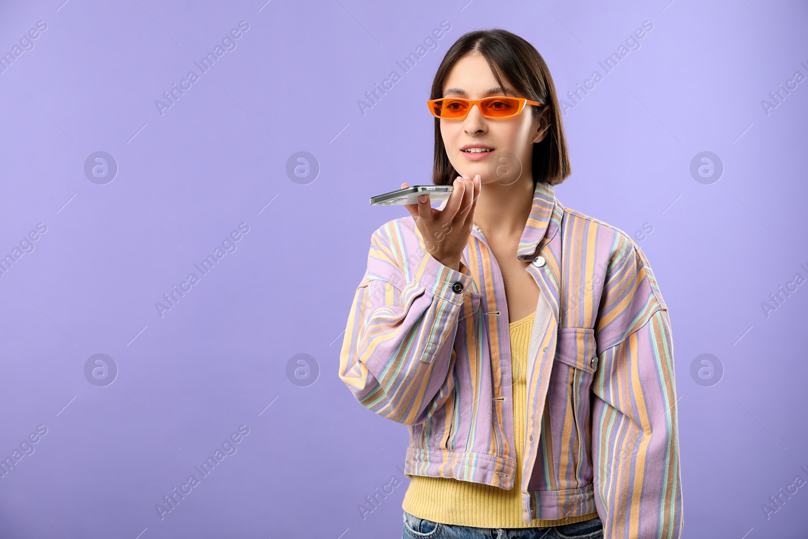 Photo of Beautiful woman in sunglasses recording voice message via smartphone on violet background, space for text