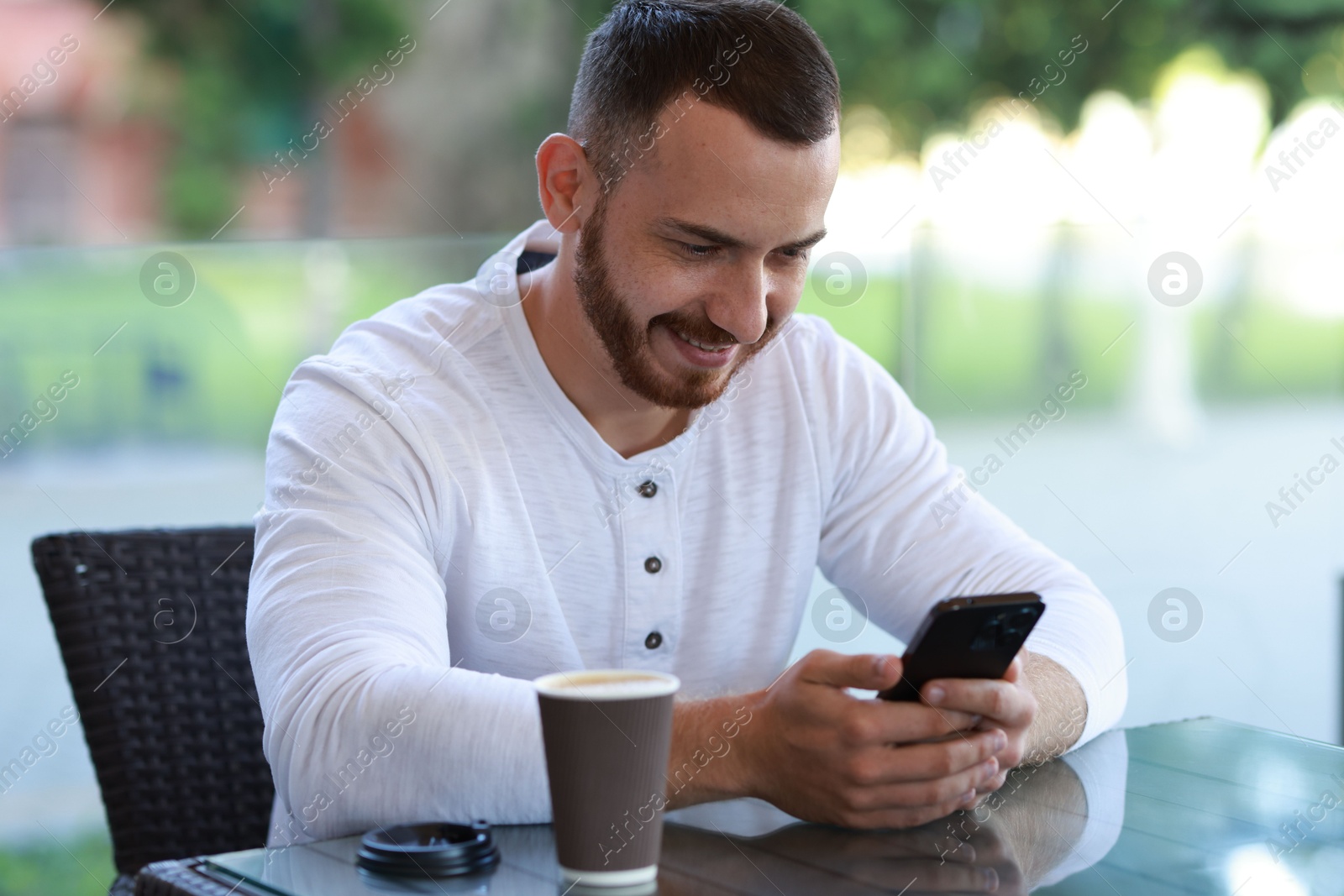 Photo of Happy man with paper cup using smartphone at outdoor cafe