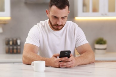 Handsome man using smartphone at white table in kitchen