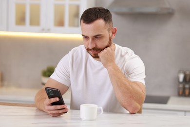 Handsome man using smartphone at white table in kitchen