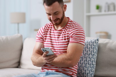 Happy man using smartphone on sofa indoors