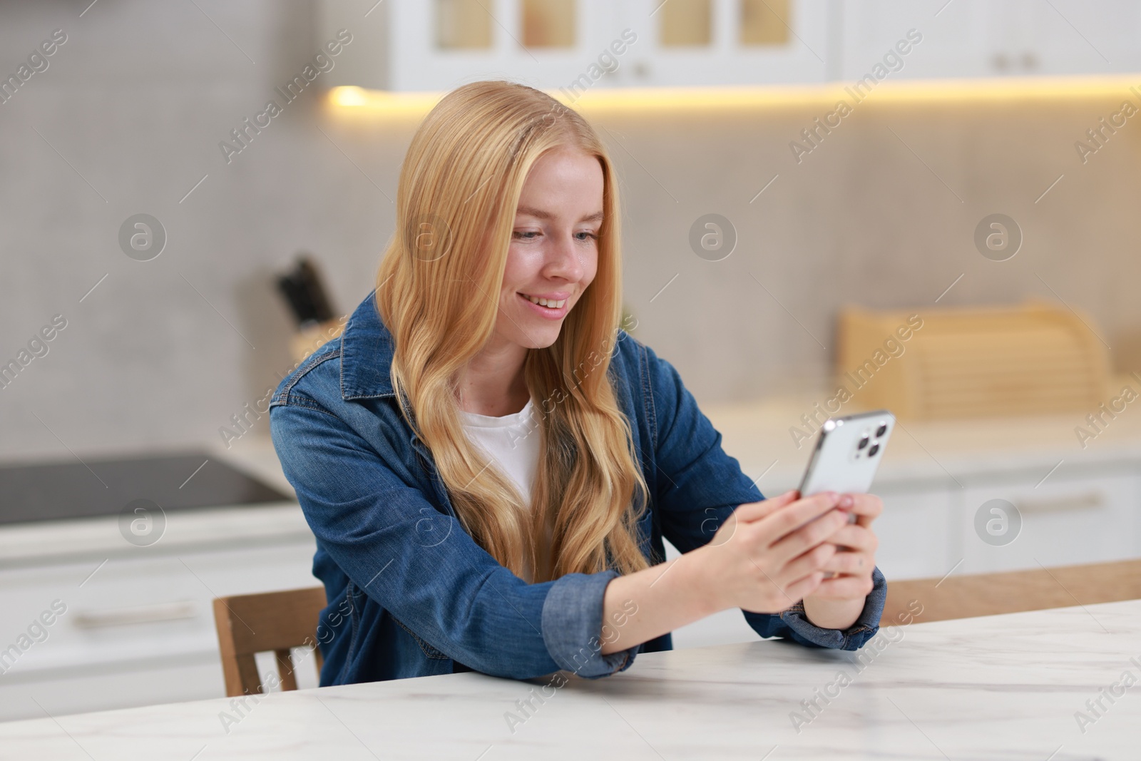 Photo of Smiling woman using smartphone at white table in kitchen