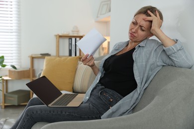 Menopause. Woman waving paper sheet to cool herself during hot flash on sofa at home