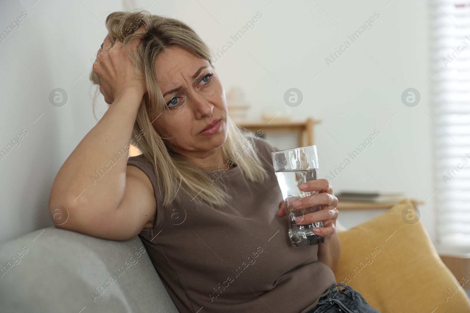 Photo of Menopause. Woman with glass of water suffering from headache on sofa at home