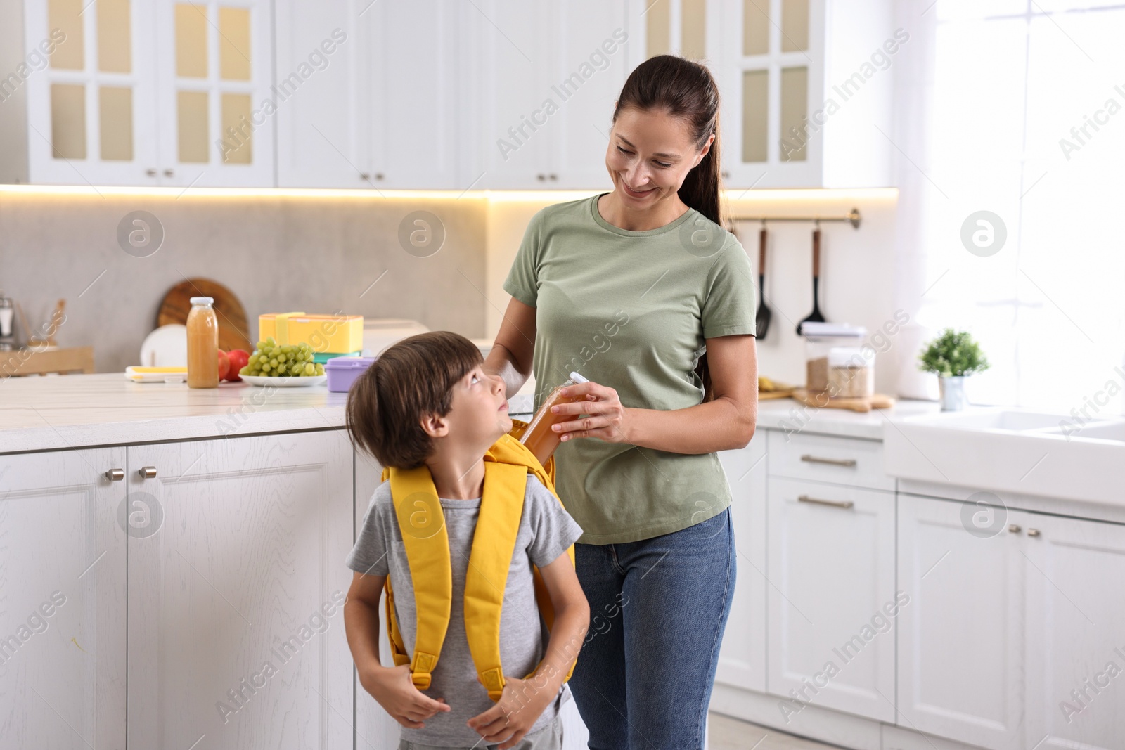 Photo of Smiling mother putting bottle of drink into son`s backpack in kitchen. Preparing for school