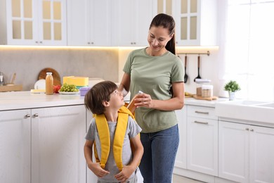 Smiling mother putting bottle of drink into son`s backpack in kitchen. Preparing for school