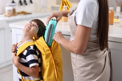 Photo of Mother putting lunch box into son`s backpack in kitchen, closeup. Preparing for school