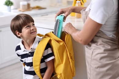 Photo of Mother putting lunch box into son`s backpack in kitchen, closeup. Preparing for school