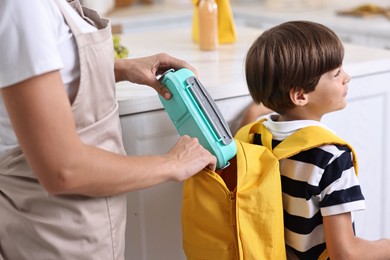 Photo of Mother putting lunch box into son`s backpack in kitchen, closeup. Preparing for school