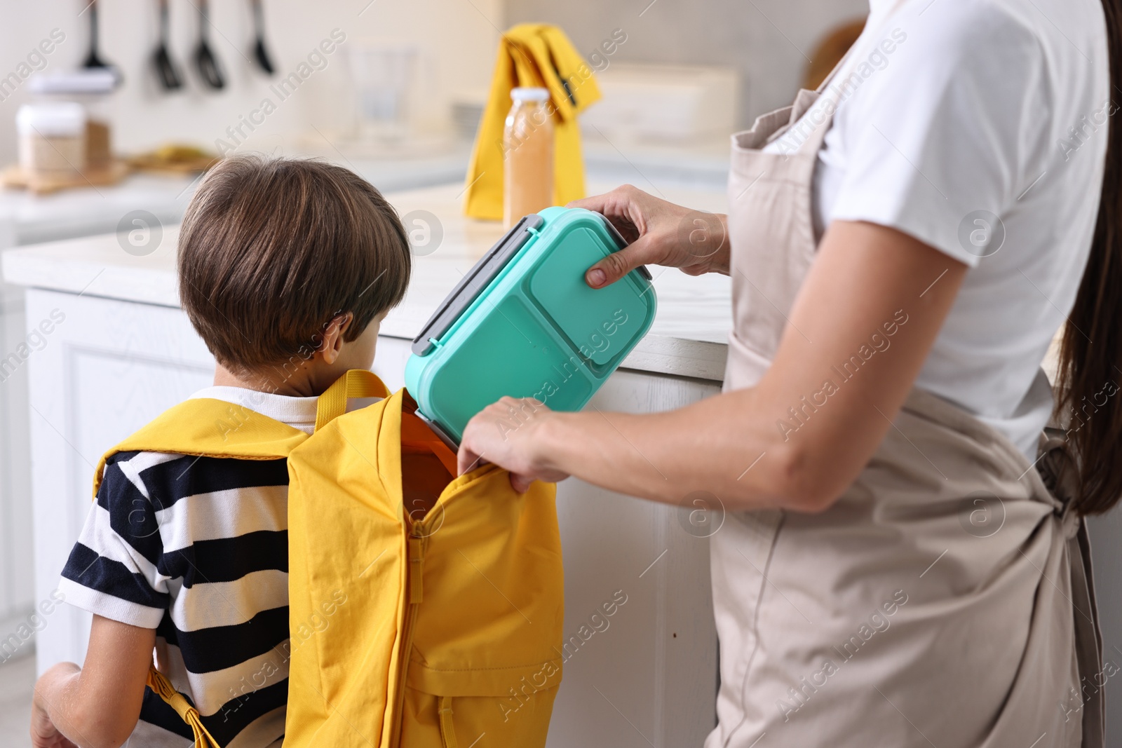 Photo of Mother putting lunch box into son`s backpack in kitchen, closeup. Preparing for school