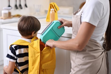 Photo of Mother putting lunch box into son`s backpack in kitchen, closeup. Preparing for school