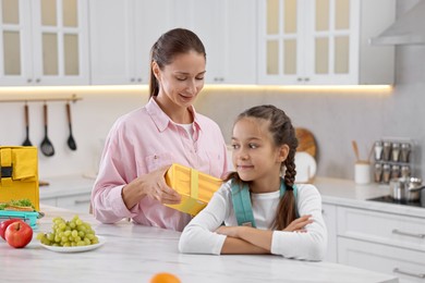 Smiling mother putting lunch box into daughter`s backpack in kitchen. Preparing for school