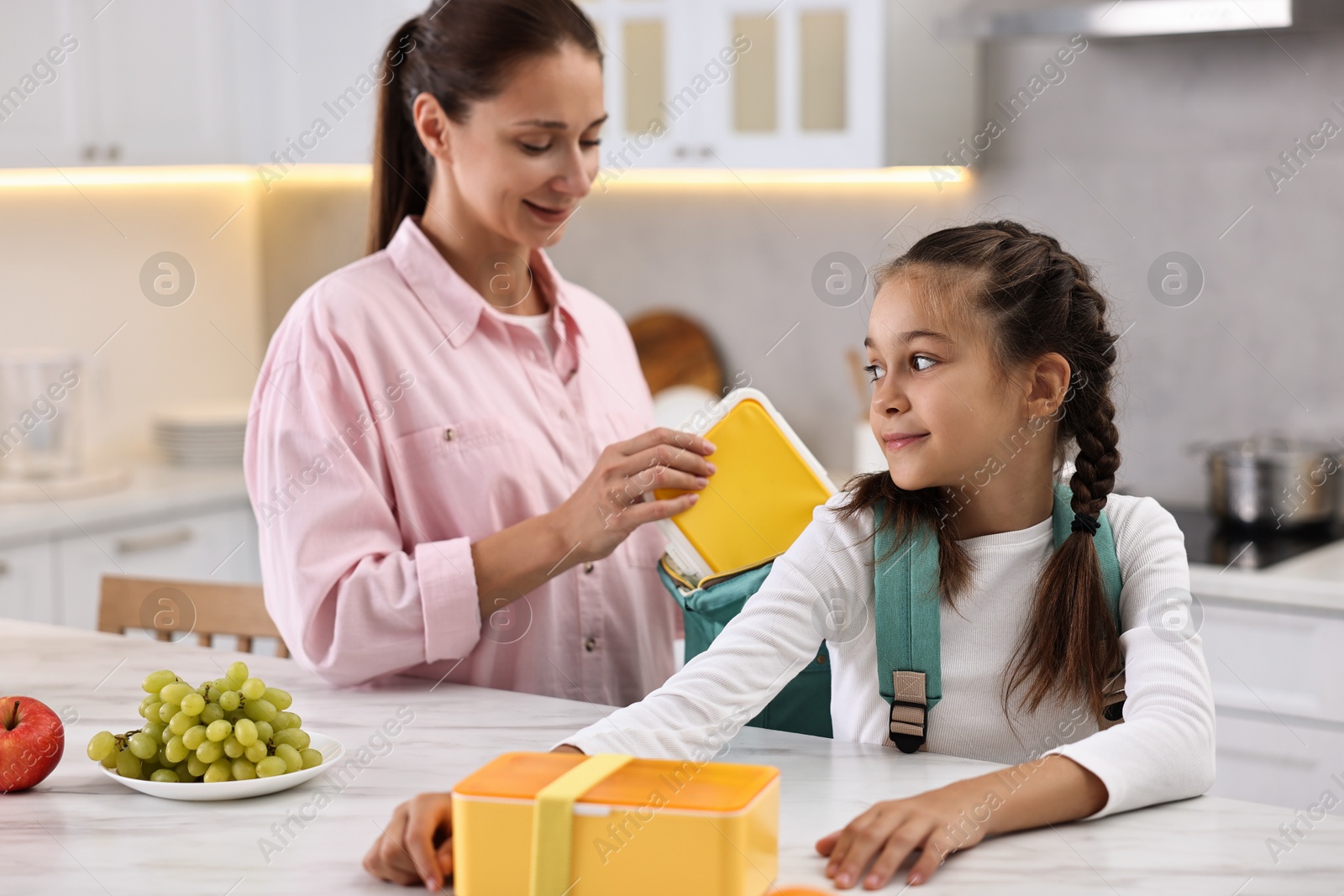 Photo of Smiling mother putting lunch box into daughter`s backpack in kitchen. Preparing for school