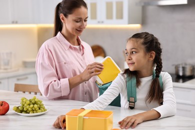 Smiling mother putting lunch box into daughter`s backpack in kitchen. Preparing for school