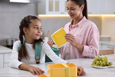 Photo of Smiling mother putting lunch box into daughter`s backpack in kitchen. Preparing for school