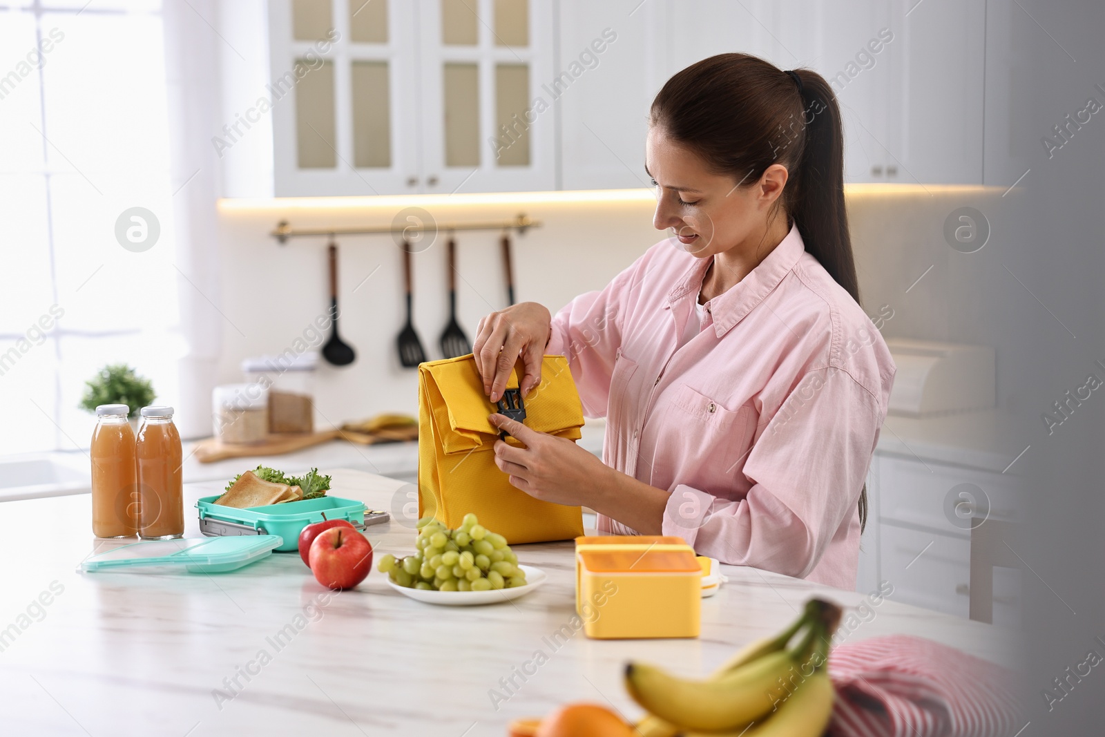 Photo of Woman with bag packing school lunch box and healthy food at white marble table in kitchen