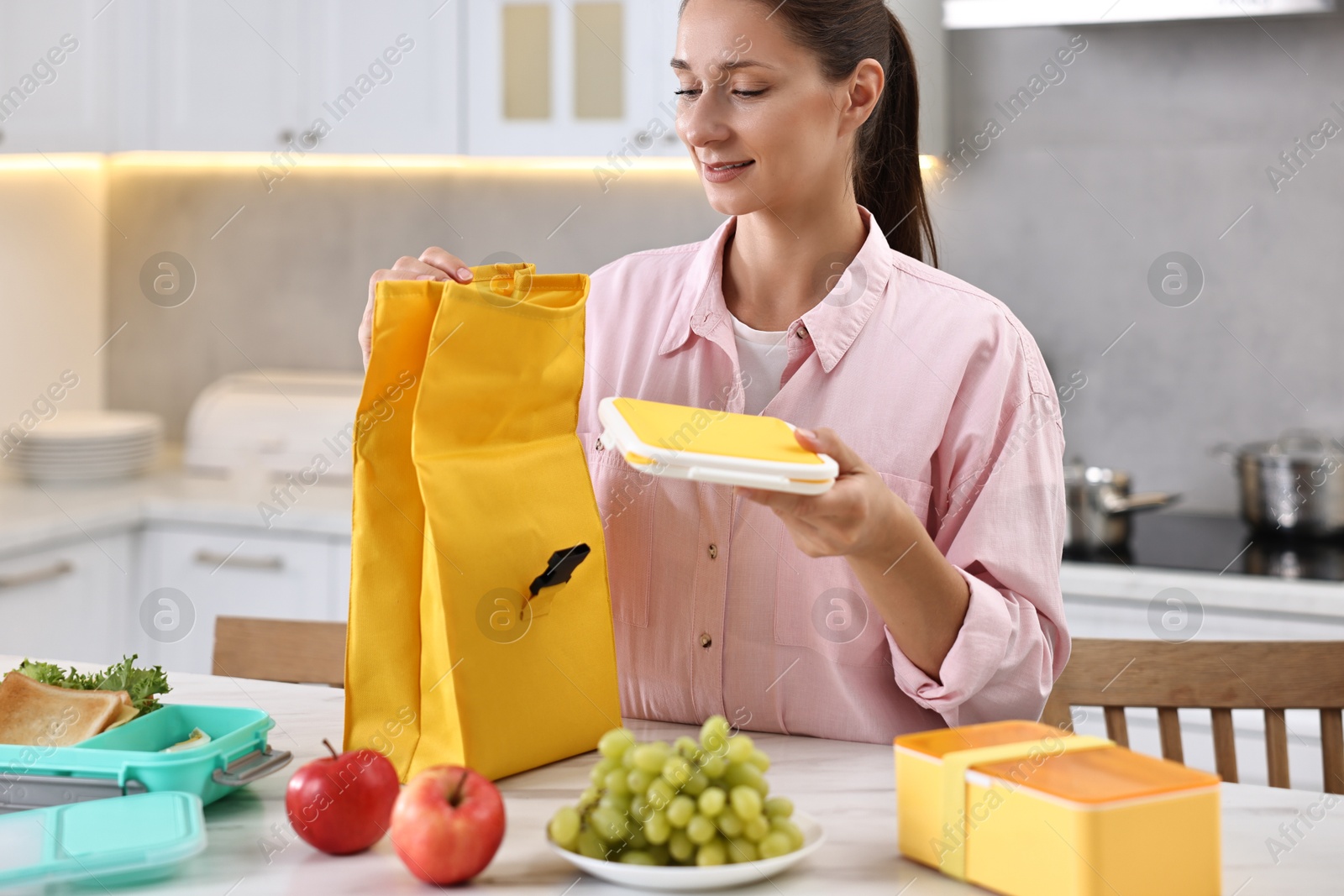 Photo of Woman with bag packing school lunch box and healthy food at white marble table in kitchen