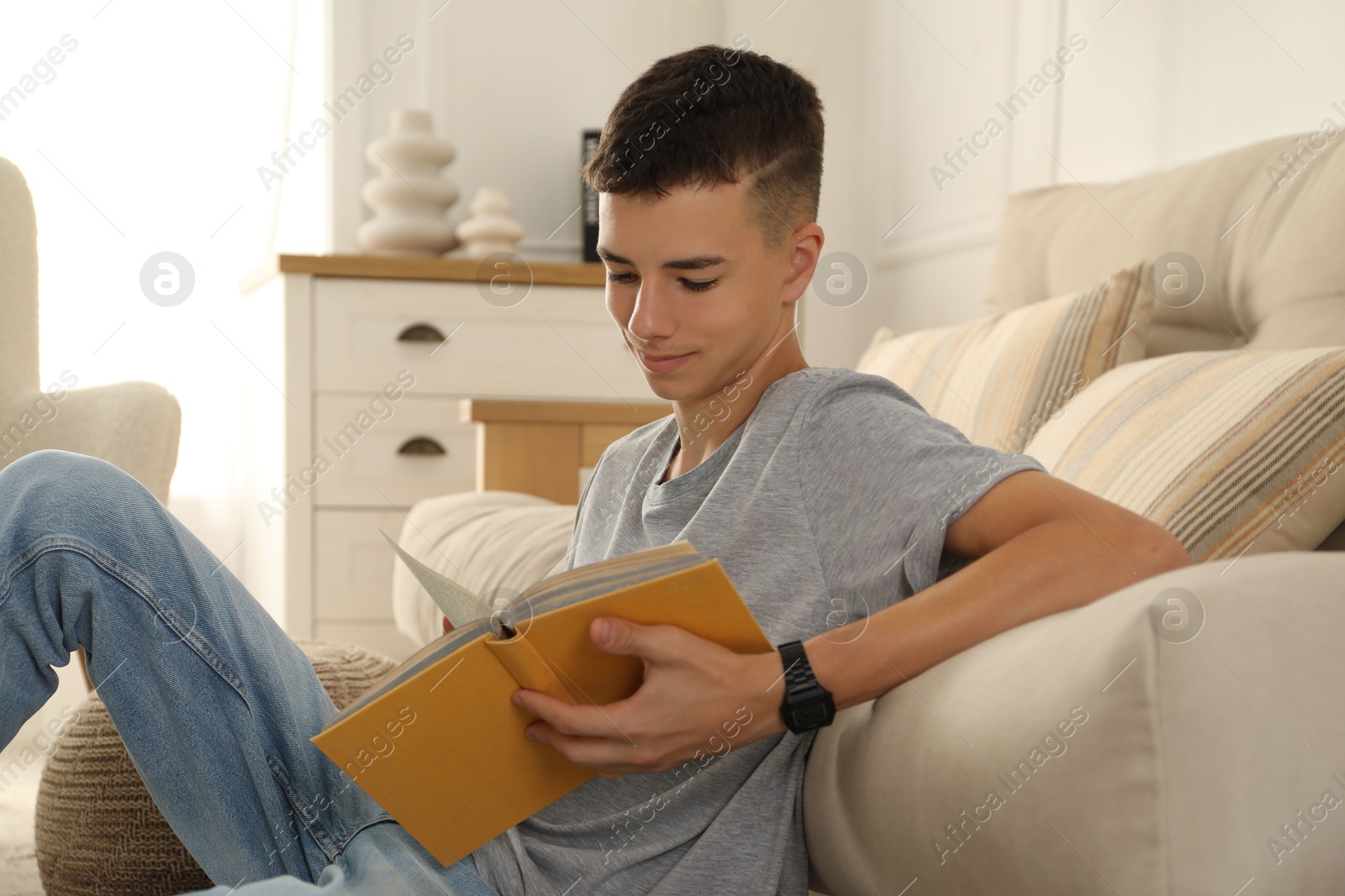 Photo of Teenage boy reading book near couch at home