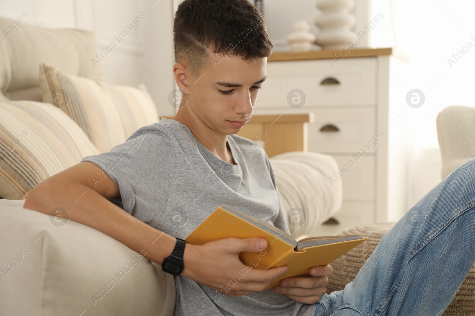 Photo of Teenage boy reading book near couch at home