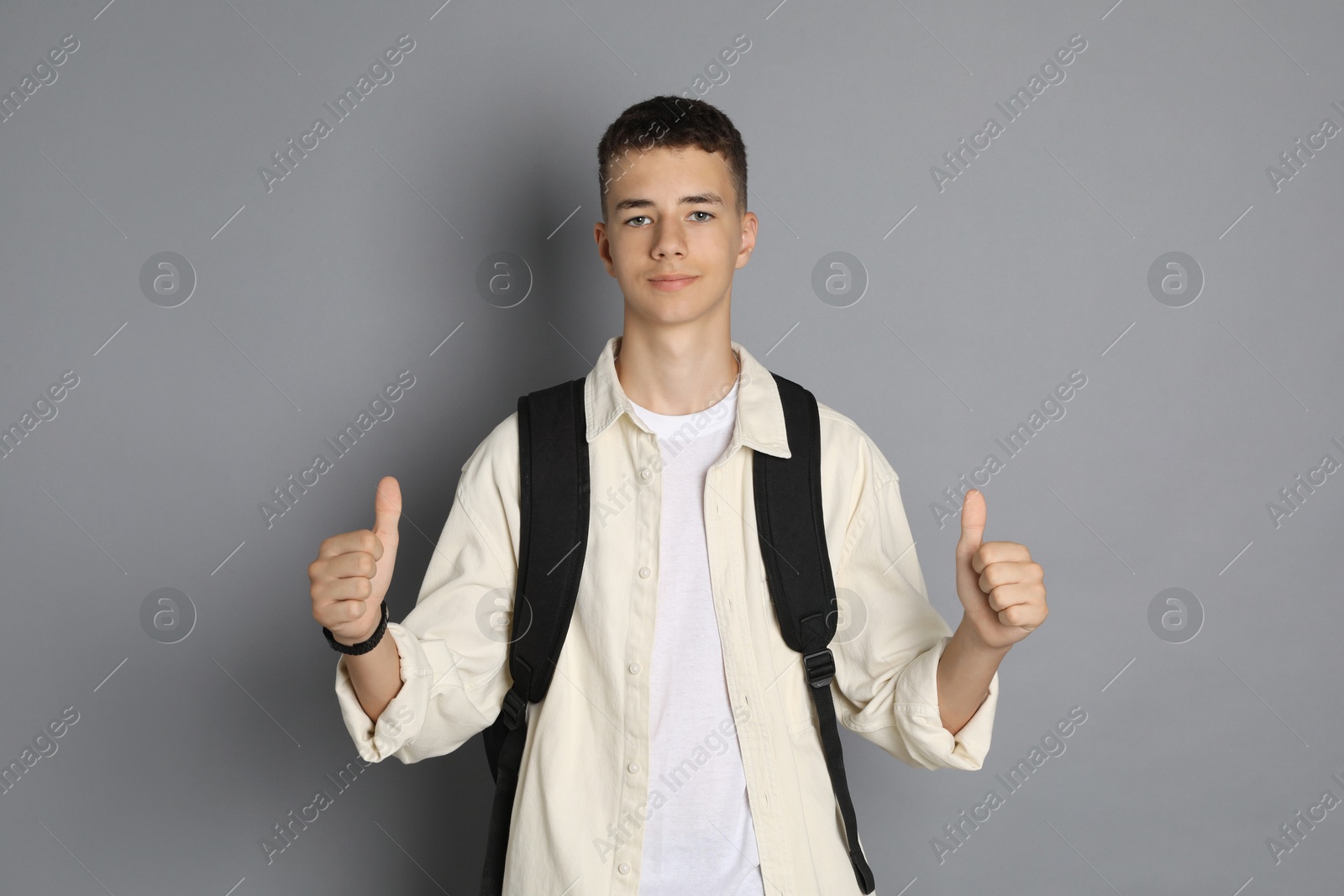 Photo of Portrait of teenage boy with backpack showing thumbs up on grey background