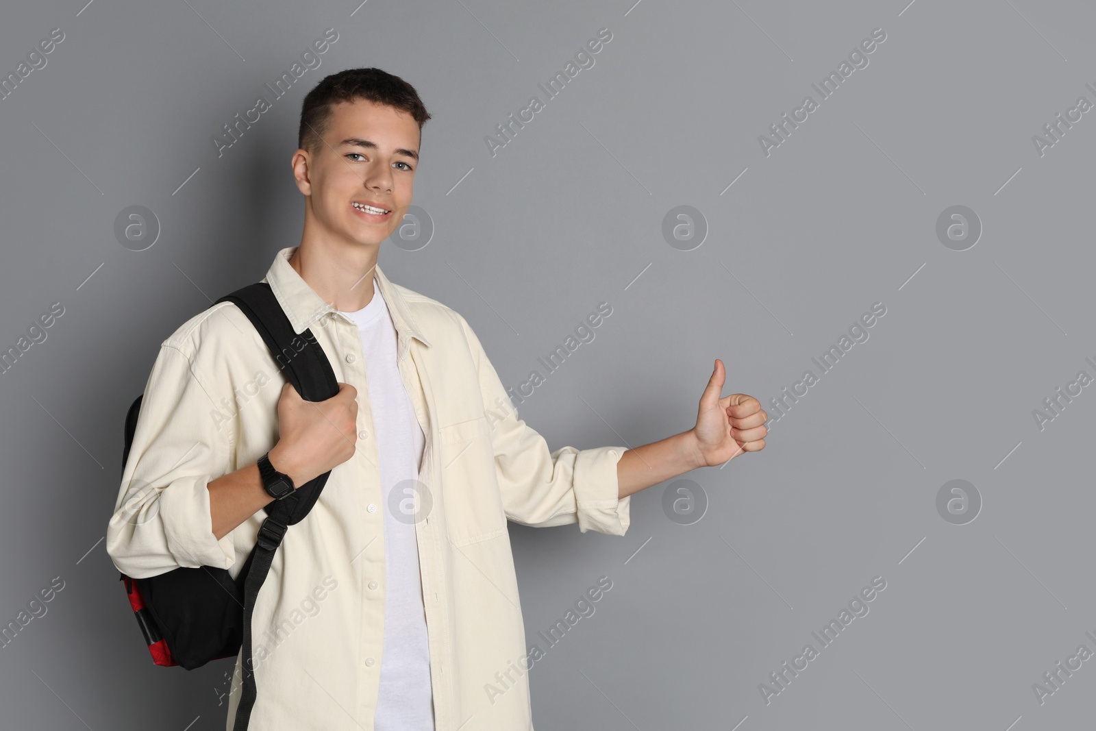 Photo of Portrait of teenage boy with backpack showing thumbs up on grey background, space for text