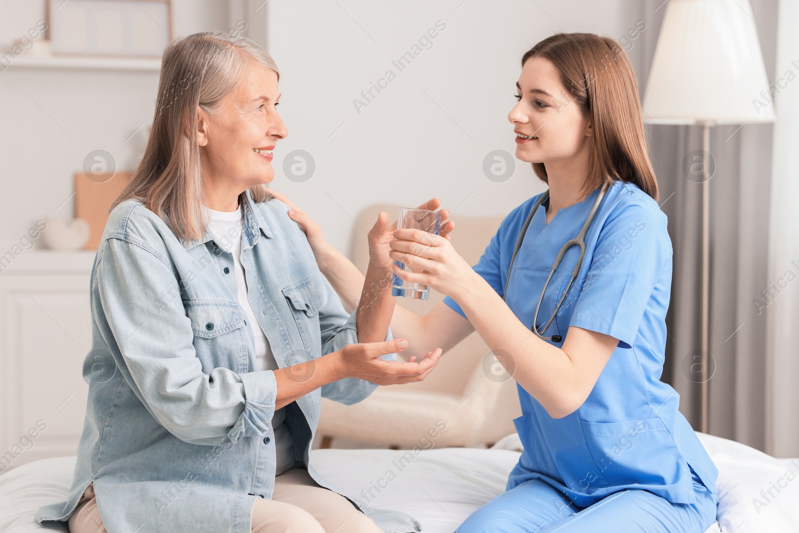 Photo of Young healthcare worker giving glass of water to senior patient on bed indoors