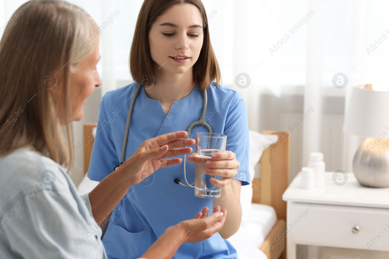 Photo of Young healthcare worker giving glass of water to senior patient on bed indoors