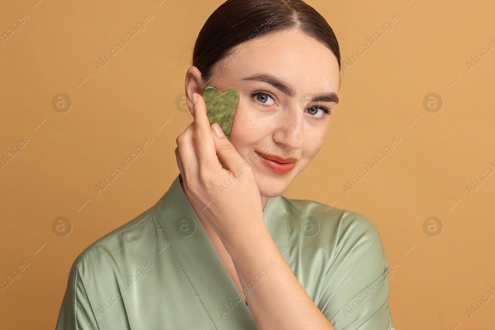 Photo of Beautiful young woman doing facial massage with gua sha tool on beige background