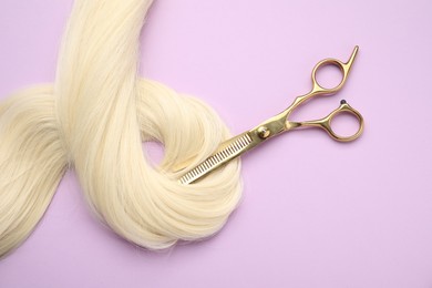 Photo of Blonde hair strand and professional scissors on violet background, top view