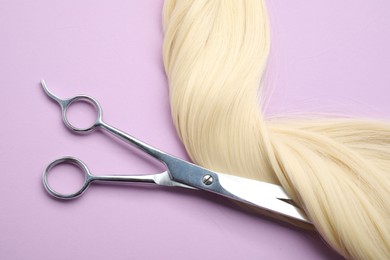 Photo of Blonde hair strand and professional scissors on violet background, top view