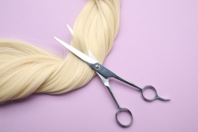 Photo of Blonde hair strand and professional scissors on violet background, top view