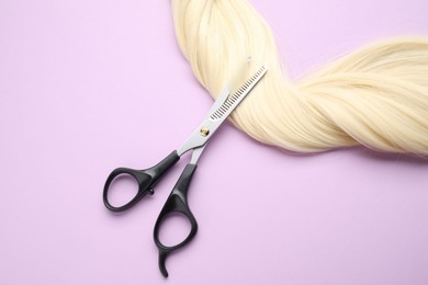 Blonde hair strand and professional scissors on violet background, top view