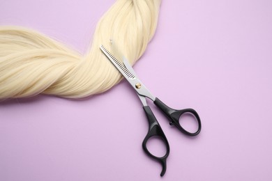 Photo of Blonde hair strand and professional scissors on violet background, top view