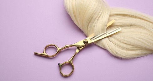 Photo of Blonde hair strand and professional scissors on violet background, top view