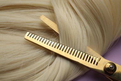 Blonde hair strand and professional scissors on violet background, top view