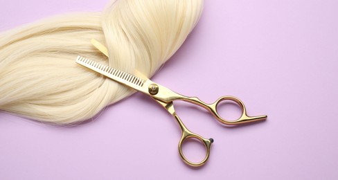 Photo of Blonde hair strand and professional scissors on violet background, top view