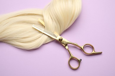 Photo of Blonde hair strand and professional scissors on violet background, top view