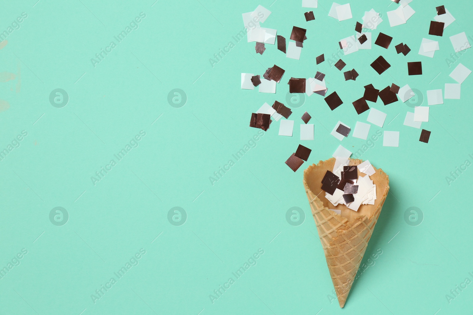 Photo of Shiny golden confetti flying out of ice cream cone on turquoise background, top view. Space for text