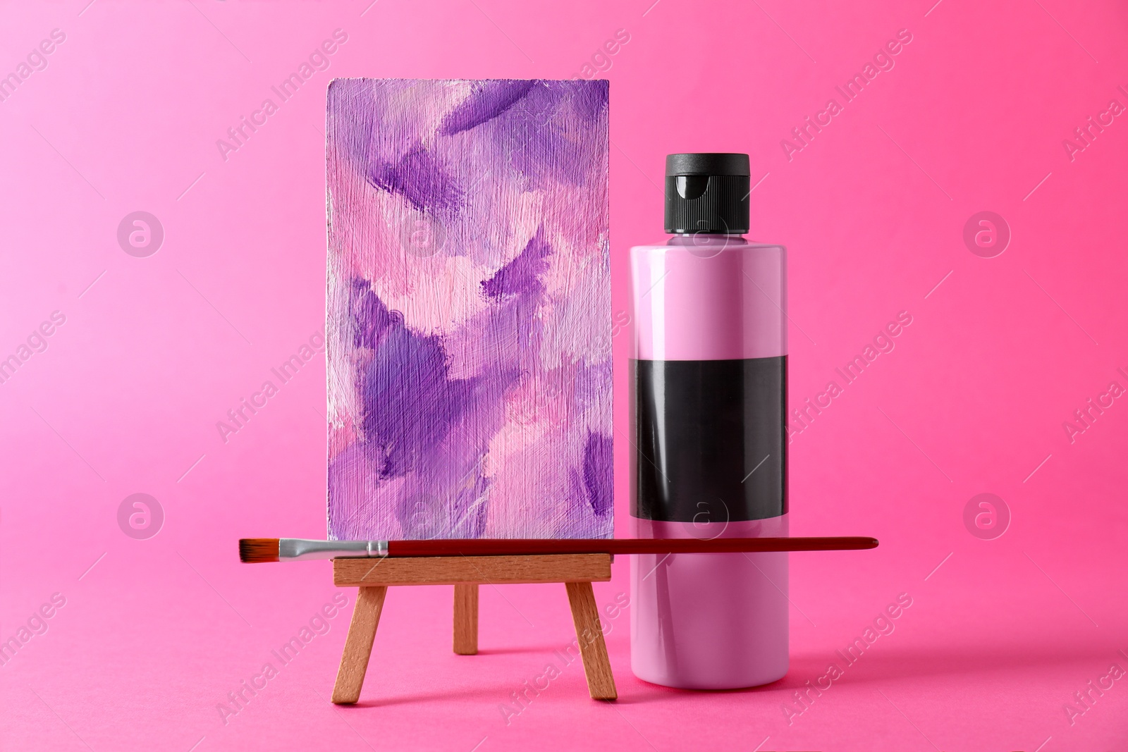 Photo of Acrylic paint in bottle, small easel with abstract picture and brush on pink background