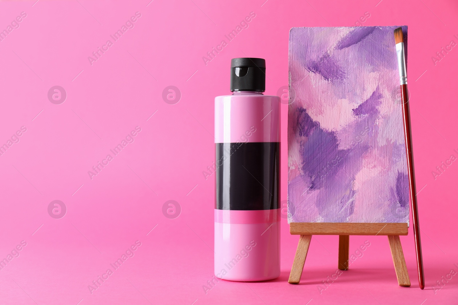 Photo of Acrylic paint in bottle, small easel with abstract picture and brush on pink background, space for text