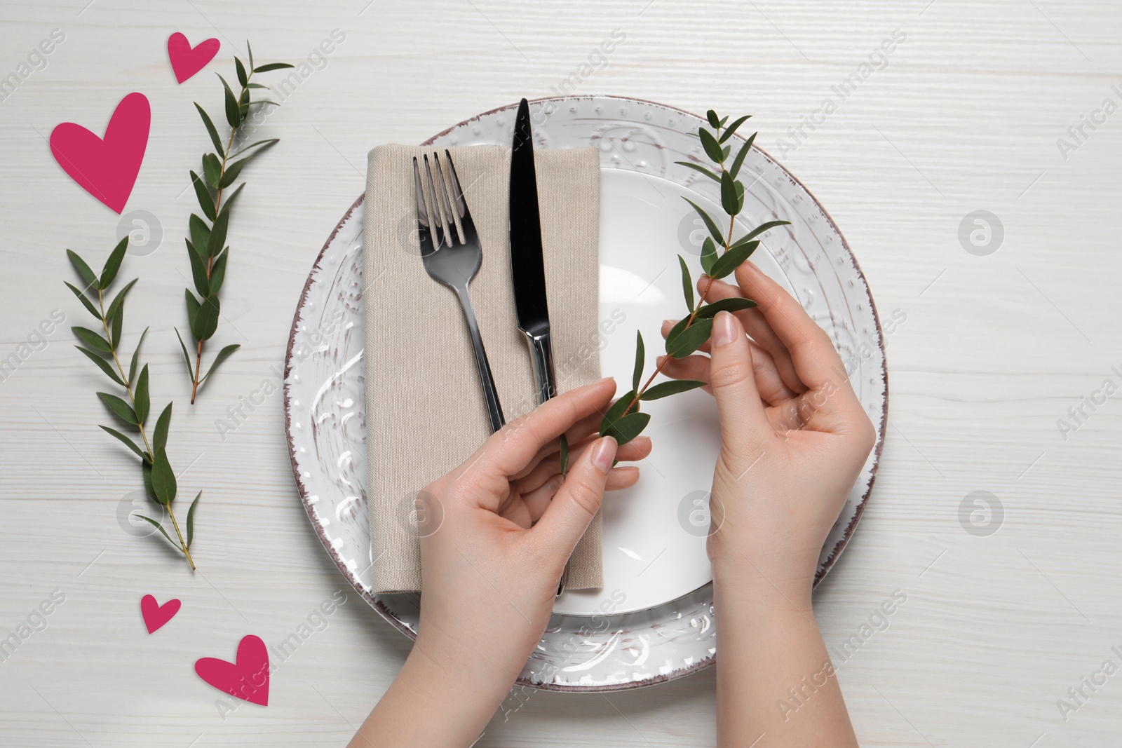 Image of Woman decorating plate with eucalyptus branches for romantic dinner at white wooden table, top view. Valentine's day celebration